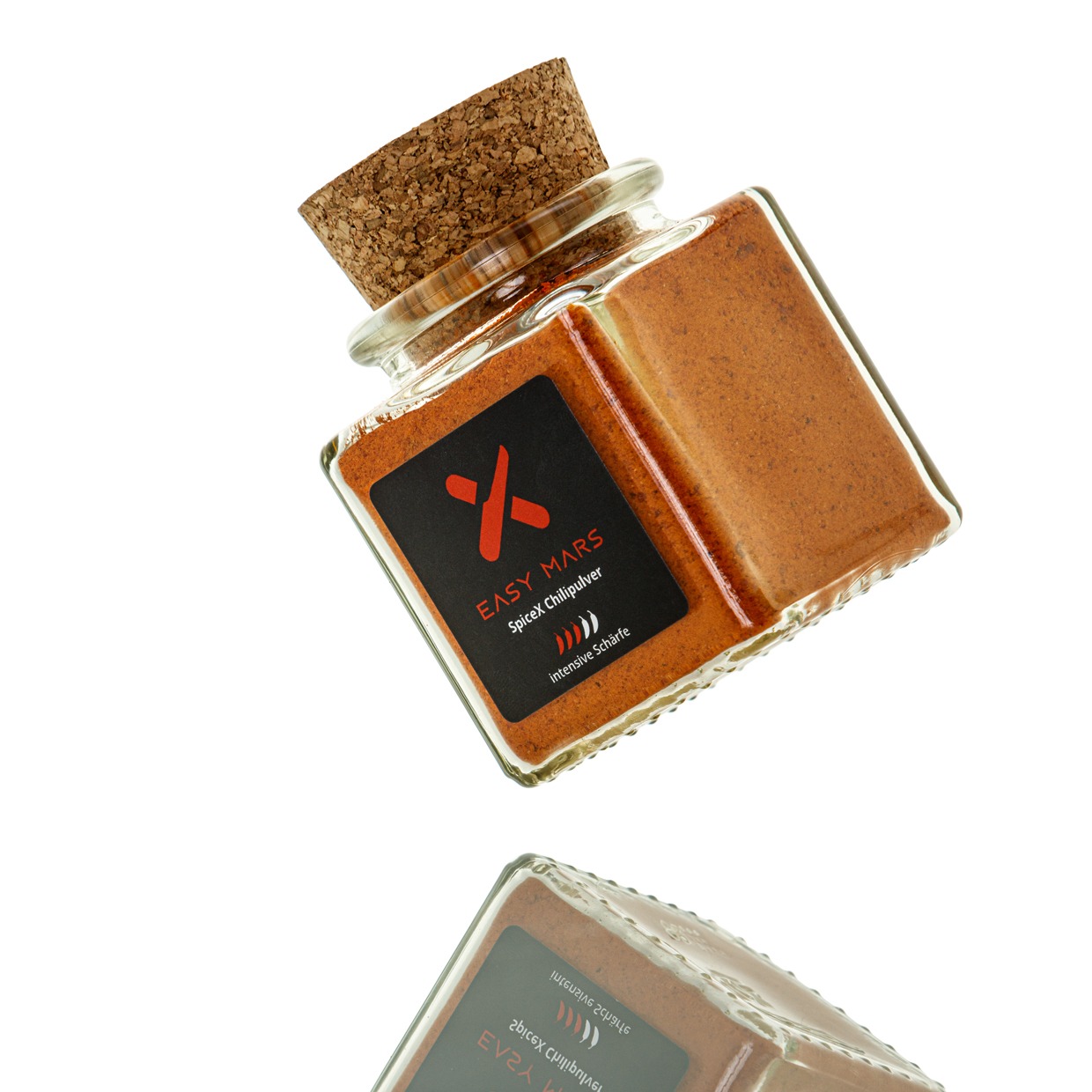 Quadratisches Glasgefäß mit Korkverschluss, gefüllt mit rotem Chilipulver, auf weißem Hintergrund freigestellt.
